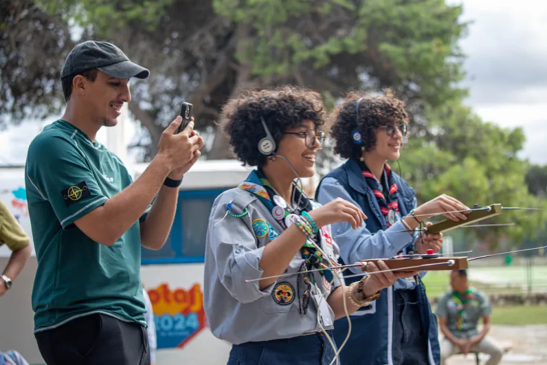 Three young scouts participating in an outdoor radio activity. Two of them are wearing headphones and holding direction-finding antennas, while a third is smiling and taking a photo with a smartphone. They appear engaged and happy. A JOTA-JOTI 2024 banner and trees are visible in the background.
